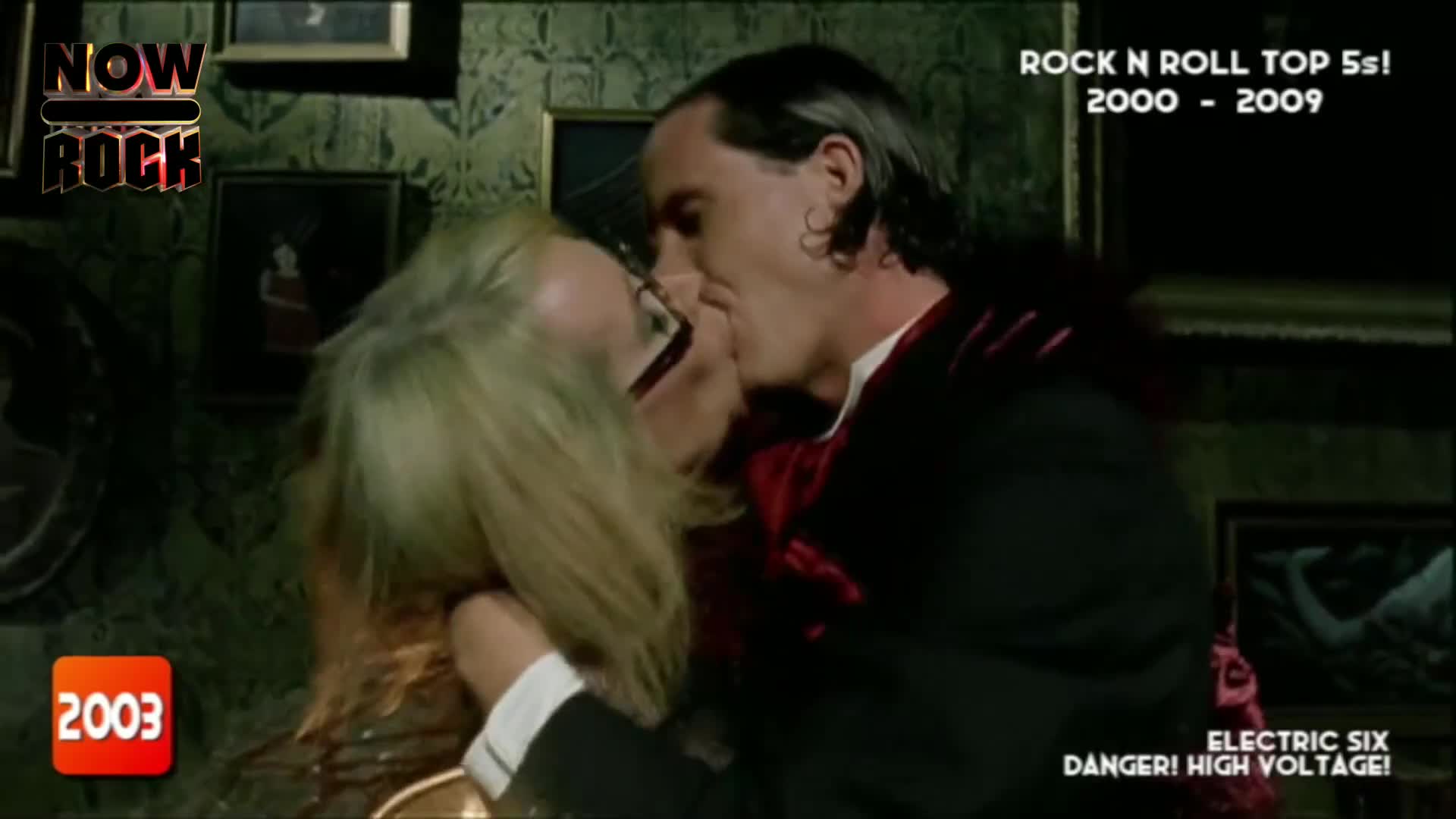 A man in a black suit and a woman with blonde hair are locked in a kiss. The scene looks like a music video from the early 2000s, with a "NOW Rock" logo in t...