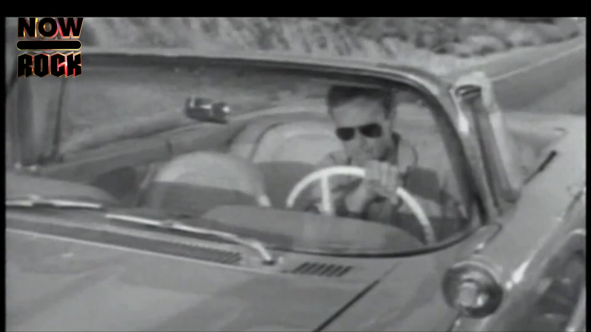 A man in sunglasses grips the white steering wheel of a classic car, heading down a road. The vehicle, a convertible, appears to be moving fast.