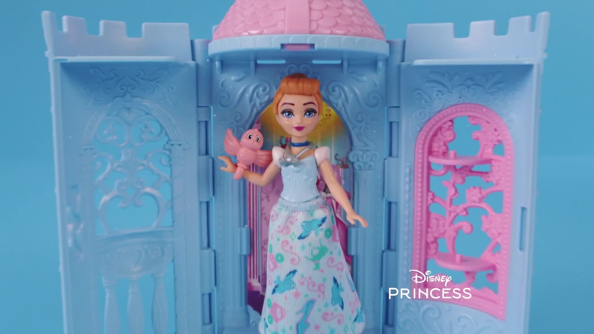 A doll, dressed in a light blue gown, stands in front of a miniature castle. She holds a small pink bird in her hand, and the Disney Princess logo is visible...