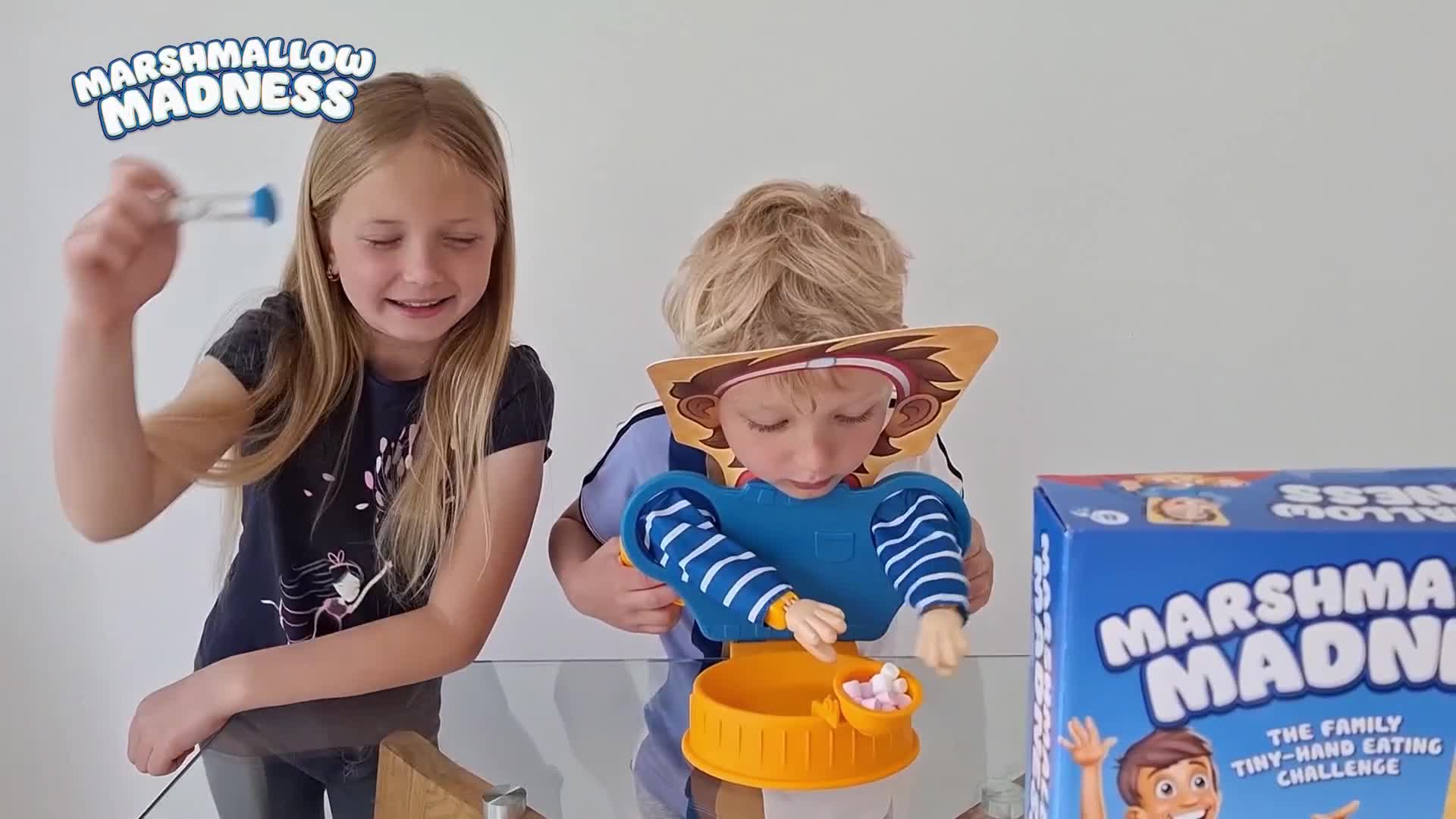 The girl is holding up a small hourglass, while the boy attempts to grab marshmallows with tiny plastic hands. A "Marshmallow Madness" game box sits on the t...