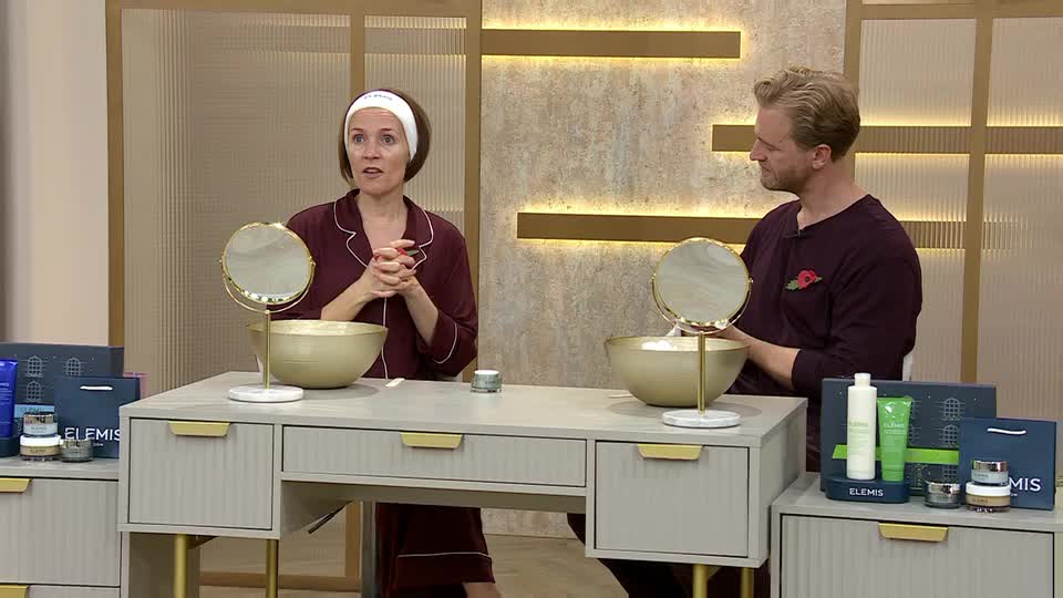 A woman in a headband and pajamas gestures with her hands, speaking to the camera. Beside her, a man in a burgundy sweater looks on, both seated at a vanity ...