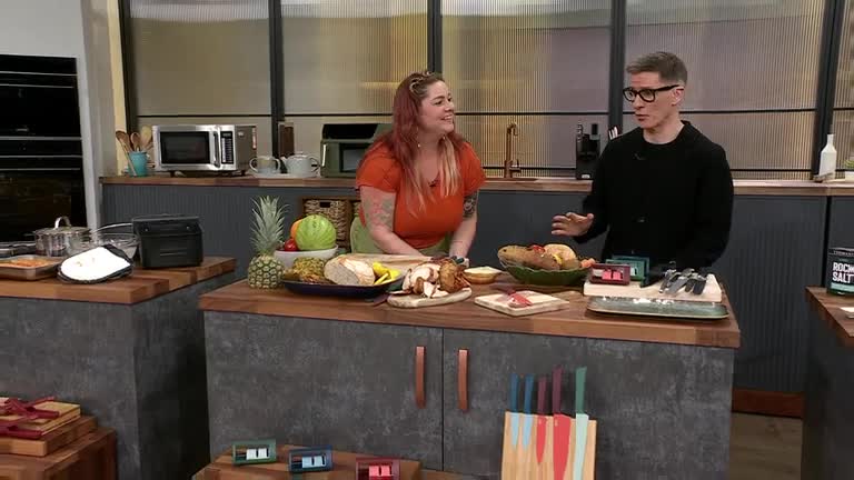 The woman in the orange top is smiling and gesturing, while the man in glasses speaks, his hand moving to emphasize a point. They're on a QVC set in the UK, ...