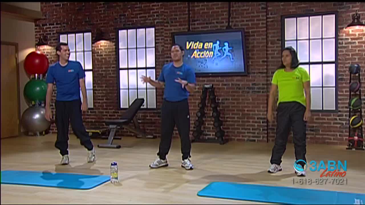 Three people are in a studio, dressed for exercise, with a "Vida en Acción" logo on a screen behind them. The man in the center gestures with his hands, whil...