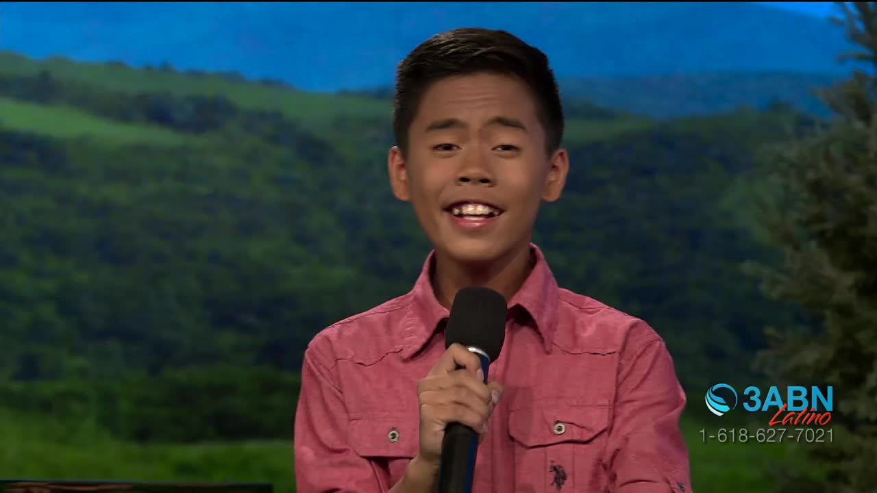 A young man in a pink shirt holds a microphone, smiling as he speaks. Behind him, a painted backdrop depicts a green landscape, and the 3ABN Latino logo is v...