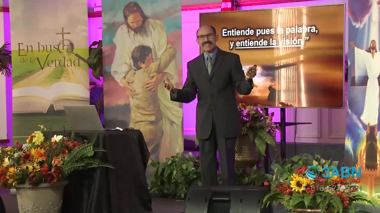 A man in a dark suit gestures with both hands, speaking in front of a screen displaying Spanish text. Behind him, colorful banners and floral arrangements de...
