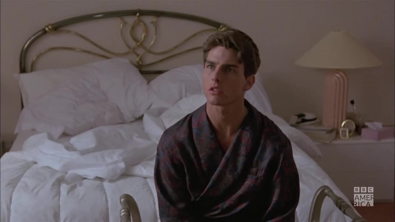 He's sitting up in bed, wearing a patterned robe, with a look of surprise on his face. The BBC America logo is visible in the corner.