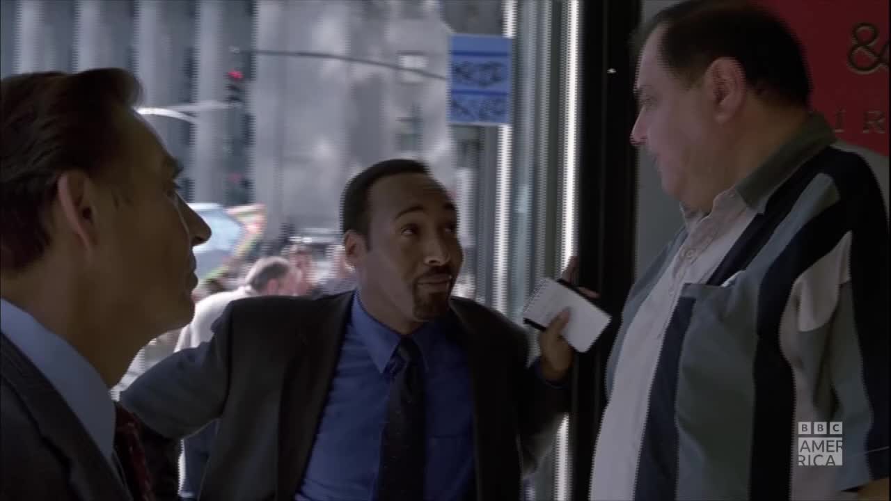 Three men stand close together, talking outside a building. The man in the middle holds a small, white card, gesturing with it as he speaks.