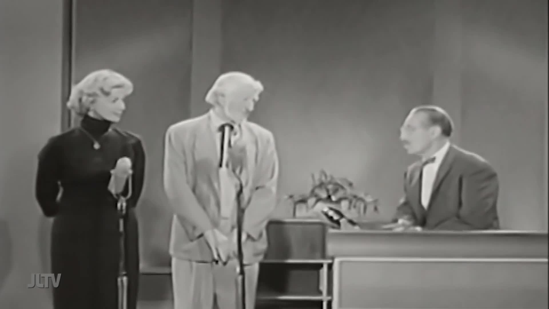 On the set of Jewish Life Television, a woman in a black dress stands next to a man in a light suit, both holding microphones. The host, in a dark suit and b...