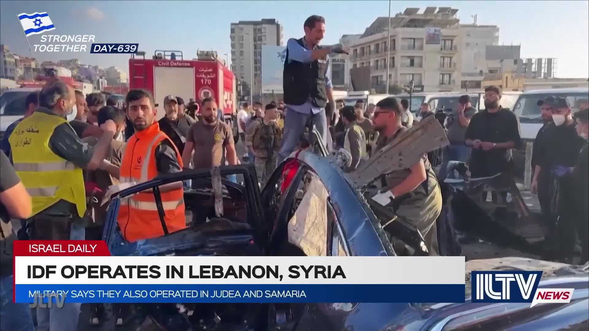 A man stands atop a wrecked car, gesturing with his hand. Rescue workers and onlookers mill around the scene, which is covered by a news banner from ILTV.