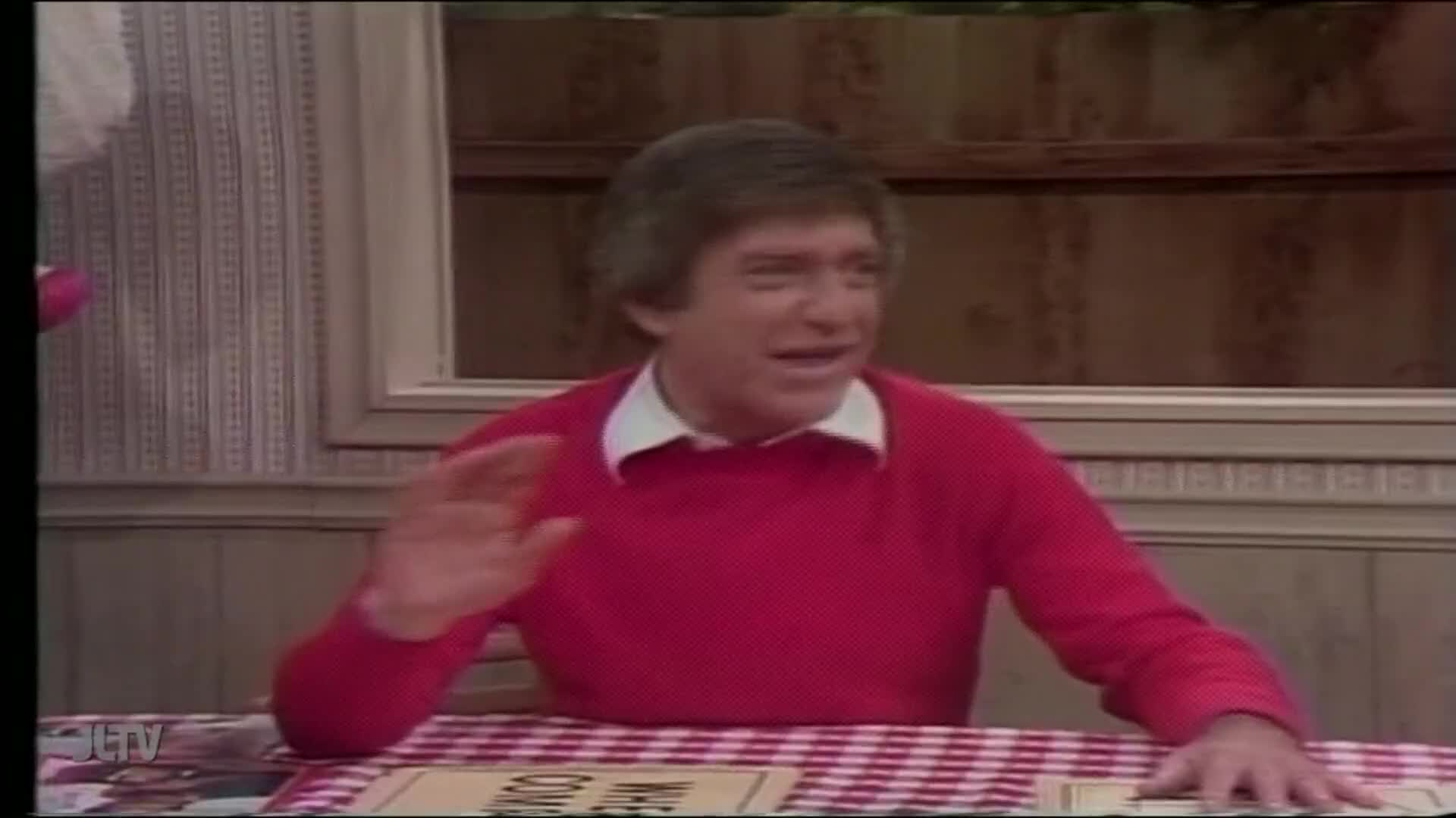 A man in a red sweater waves his hand, seated at a table with a red-and-white checkered tablecloth. The JTLV logo is visible in the corner, suggesting this i...