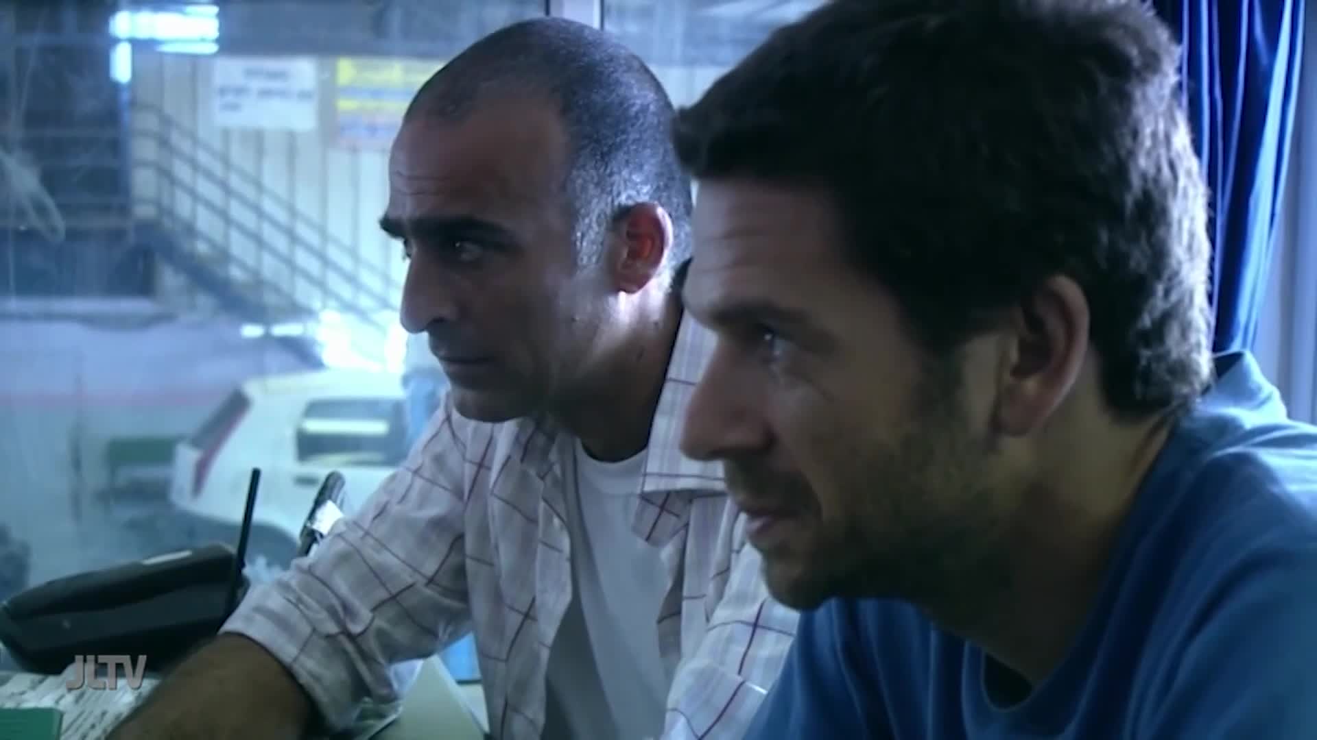 Two men, one in a checked shirt, the other in blue, are focused on something just off-screen. A "JLTV" logo is visible on a desk, and a car is parked outside...