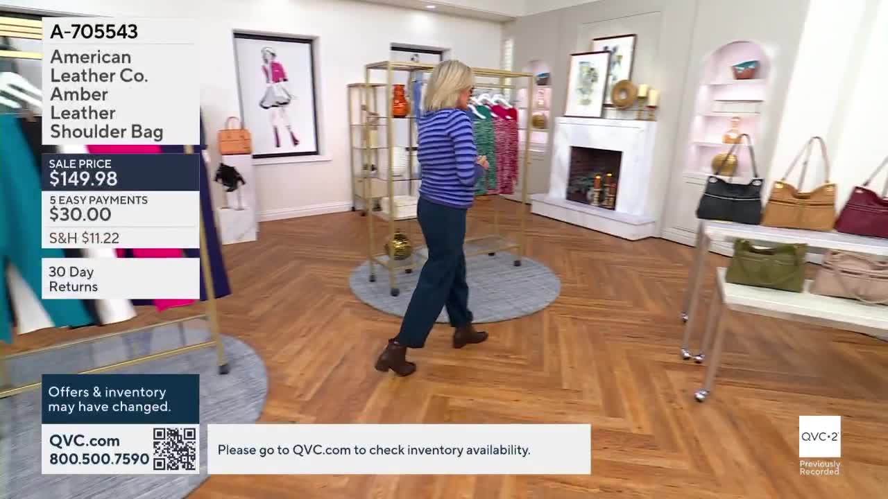 A woman in a blue striped shirt and dark pants walks across the wooden floor. The scene is from a QVC2 broadcast, with product information displayed on the l...