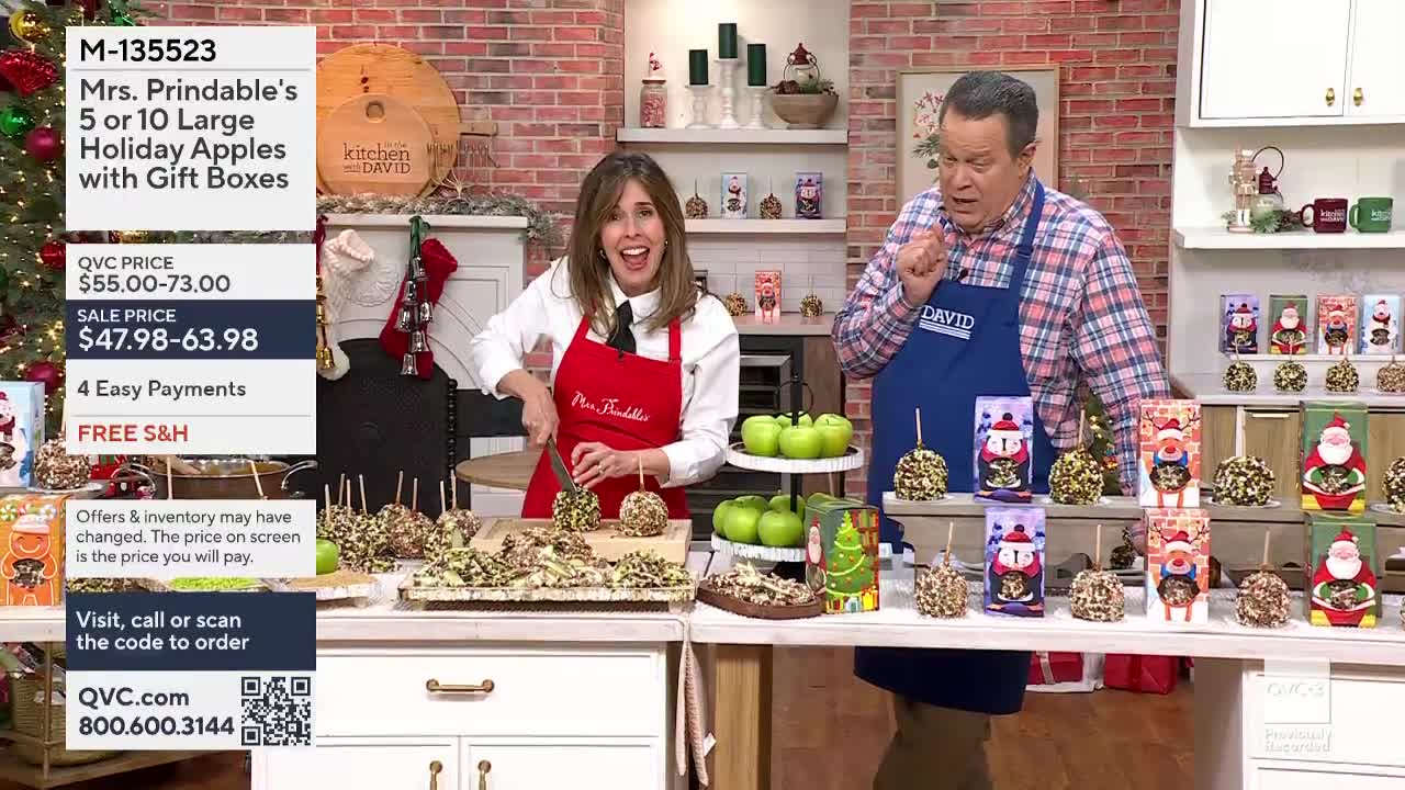 On a QVC3 set, a woman in a red apron slices a caramel apple, while a man in a blue apron gestures excitedly. Holiday-themed gift boxes and decorated apples ...