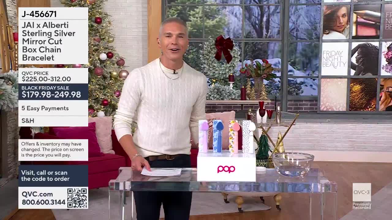 A man in a cream-colored sweater gestures towards a display of colorful items on a clear table. Behind him, a Christmas tree and a window with a winter scene...