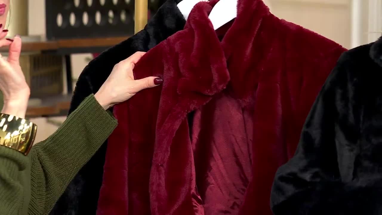 A woman's hand, with a dark red manicure, gently touches a plush, burgundy coat. Next to it, a black coat hangs on a white hanger, both on display for a QVC3...