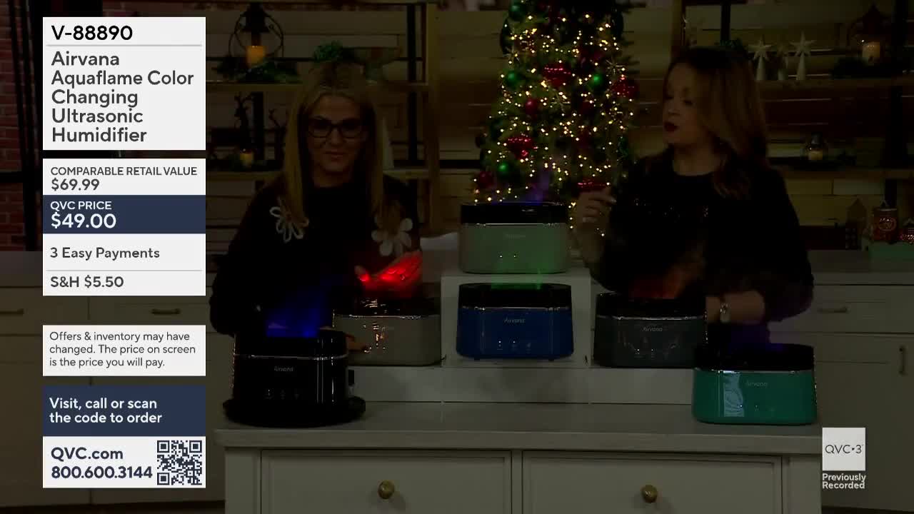 Two women stand behind a display of small humidifiers, each emitting a colored, flickering light. They're on QVC3, and one woman gestures toward the products...