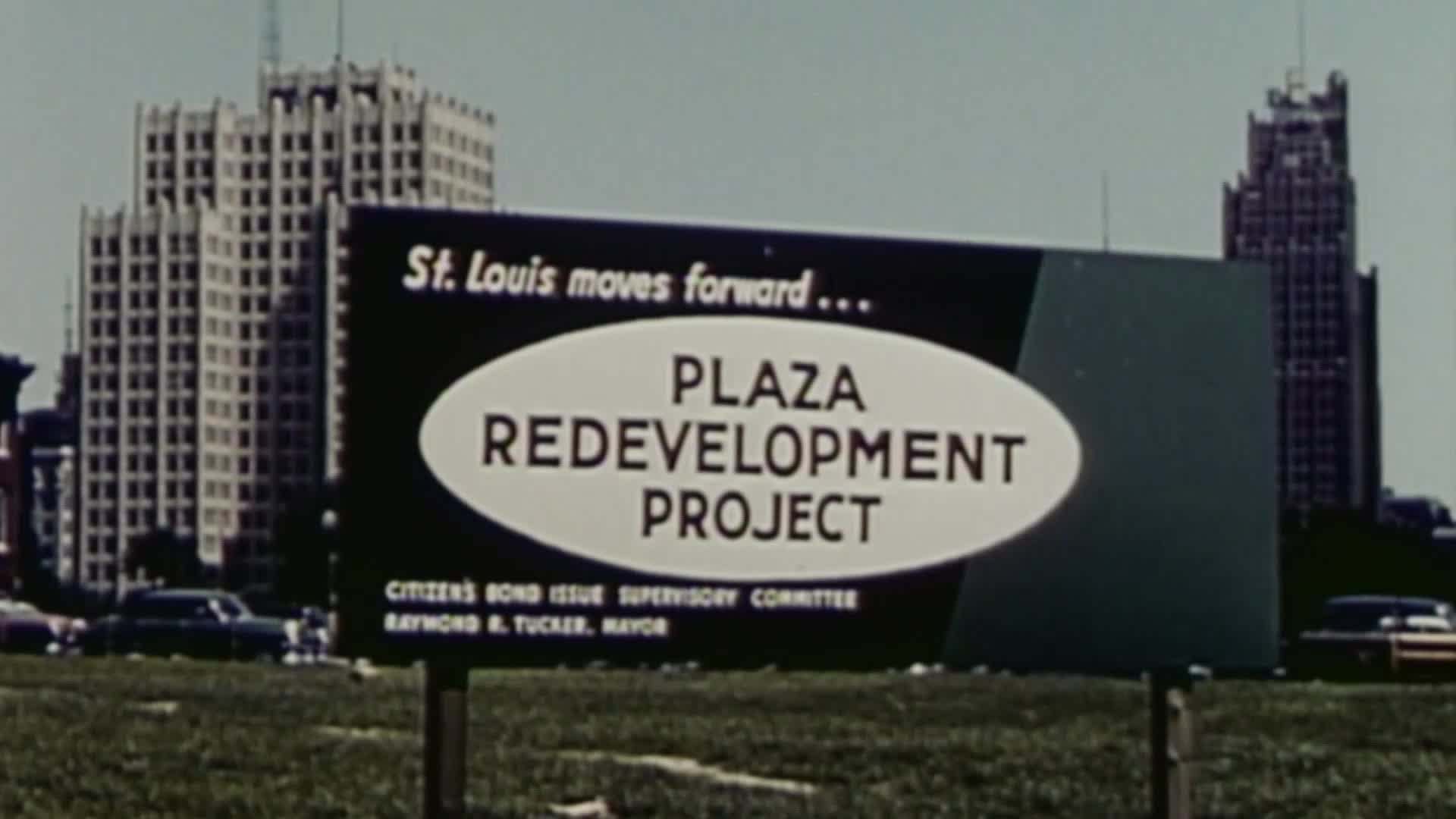 A roadside sign touts the "Plaza Redevelopment Project" in St. Louis, with a backdrop of towering buildings and a few parked cars. The sign's design and the ...