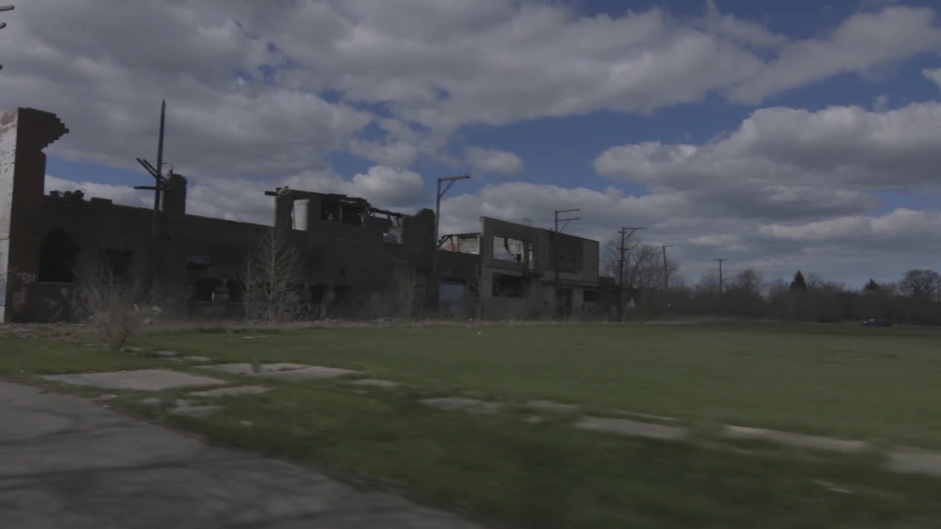 The remains of a brick building, its roof and walls partially collapsed, stand against a cloudy sky. Patches of grass and weeds cover the ground in front of ...