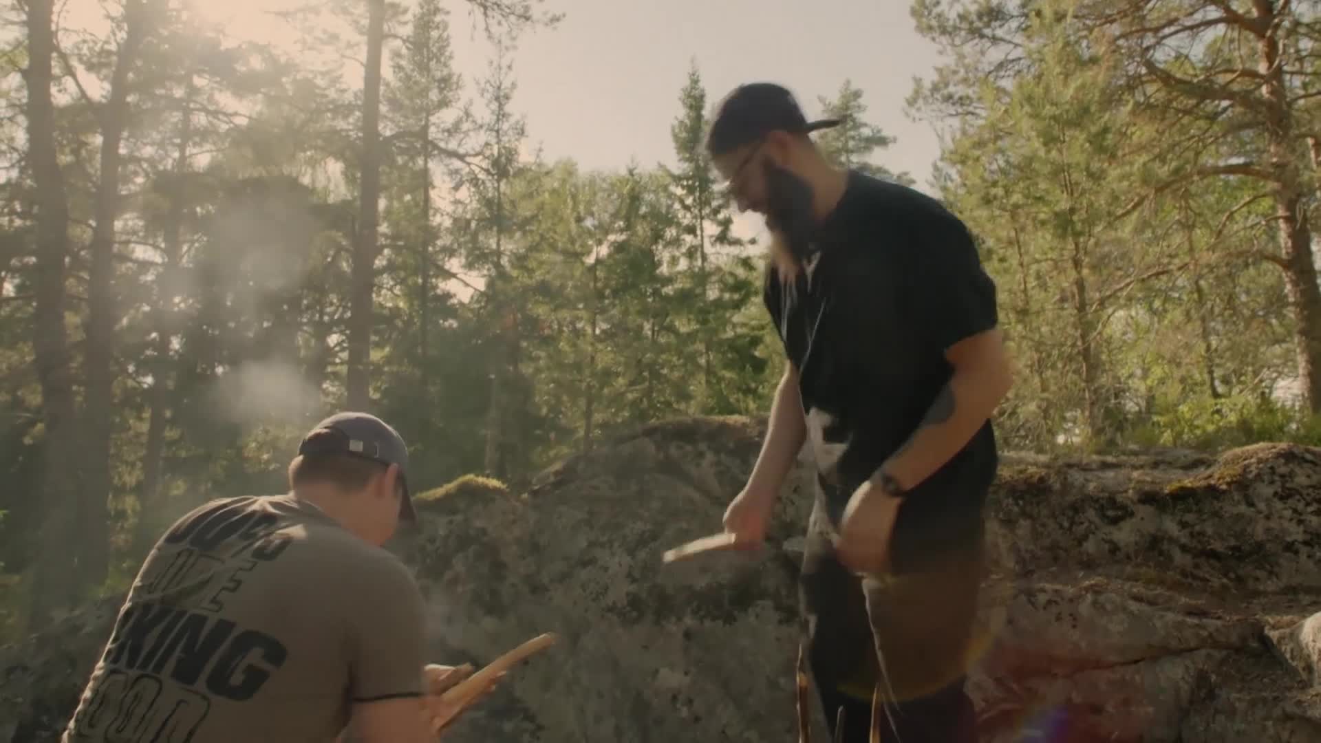 Smoke drifts up from a small fire as two men work with sticks. One, with a long beard, stands and holds a piece of wood, while the other sits, focused on his...