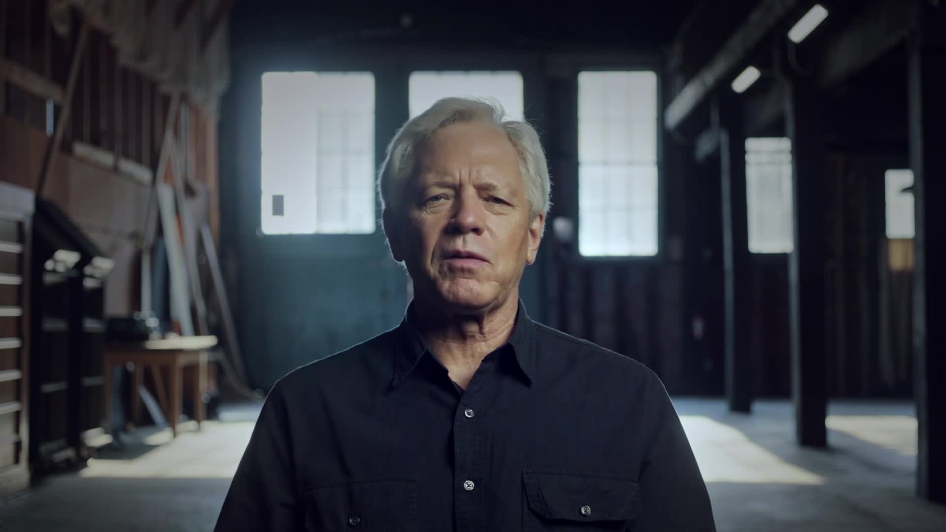 A man with silver hair and a dark shirt looks directly at the camera. He's standing in what appears to be a warehouse, with beams and windows visible in the ...