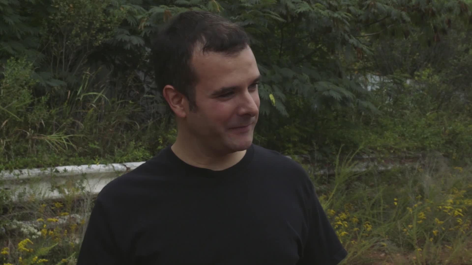 A man in a black t-shirt is looking off to the side, a slight smile on his face. He's standing in front of overgrown greenery and a white barrier.