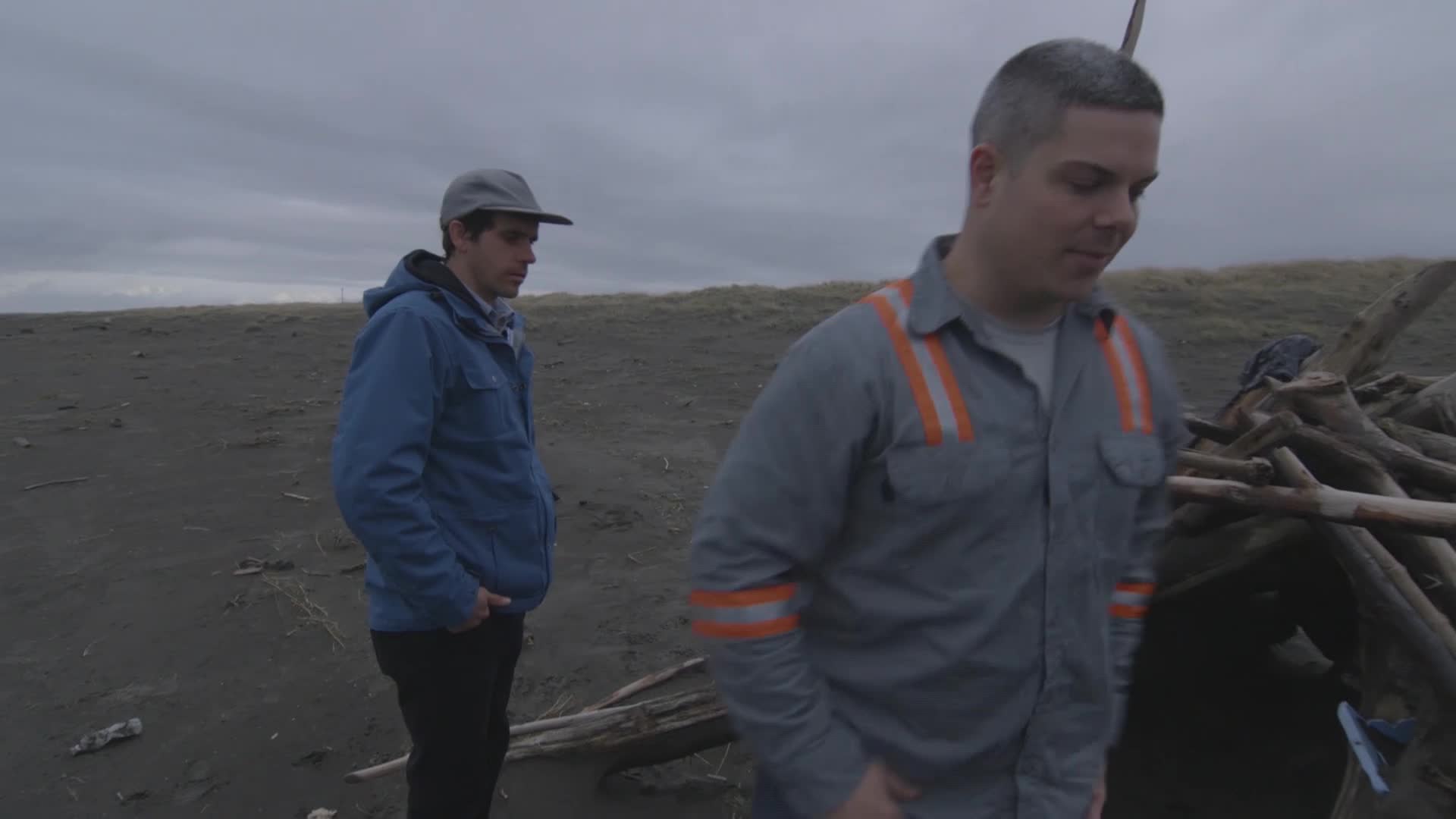 Two men stand on a dark, sandy beach under a cloudy sky. The man closest to me, wearing a work shirt with orange stripes, looks down at something out of frame.