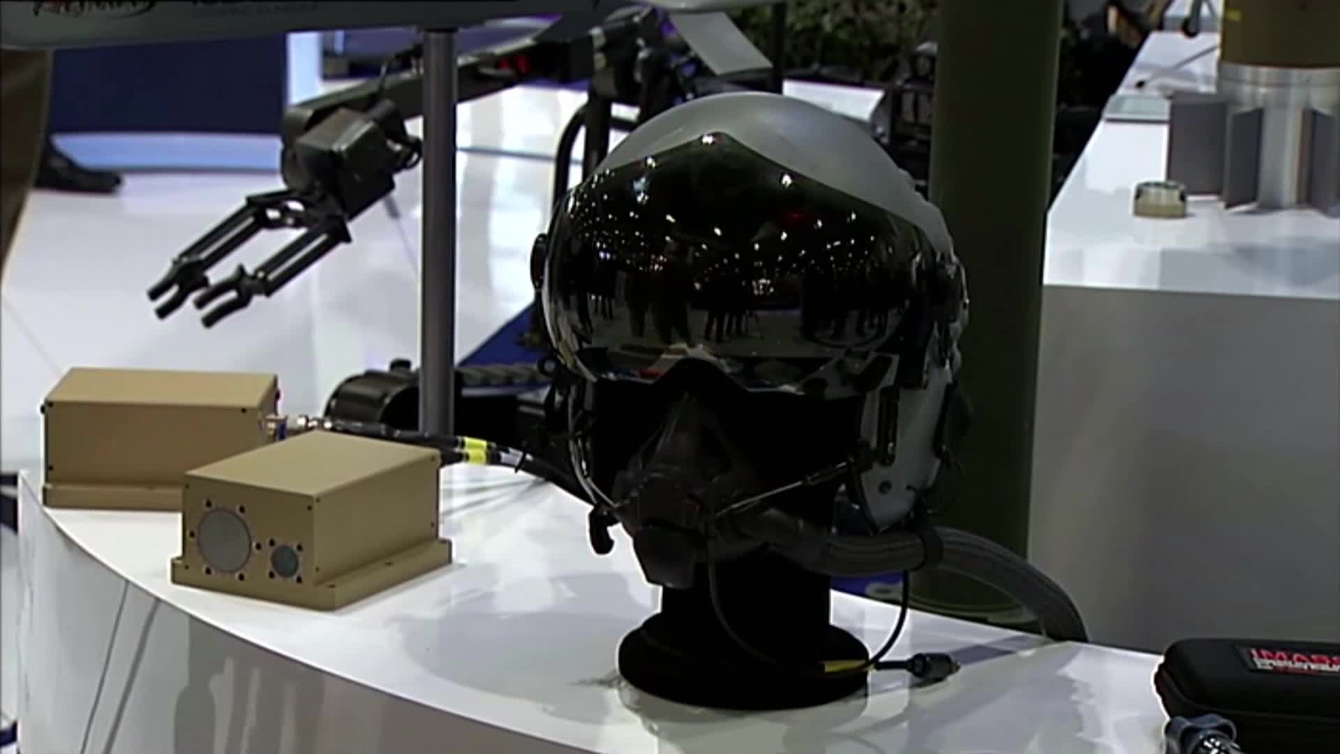 A pilot's helmet with a dark visor sits on a black stand, reflecting the surrounding environment. Two beige boxes and a robotic arm are displayed nearby, lik...