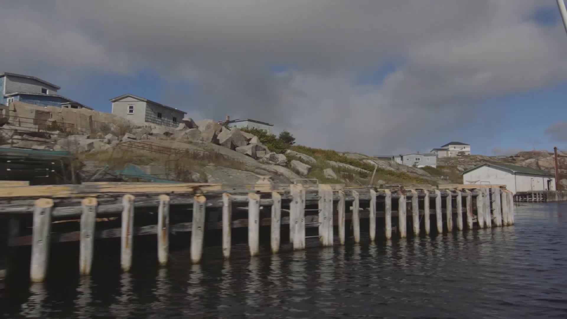 The weathered pilings of a dock stretch out into the dark water. Small, colorful houses sit perched on a rocky hillside under a cloudy sky.
