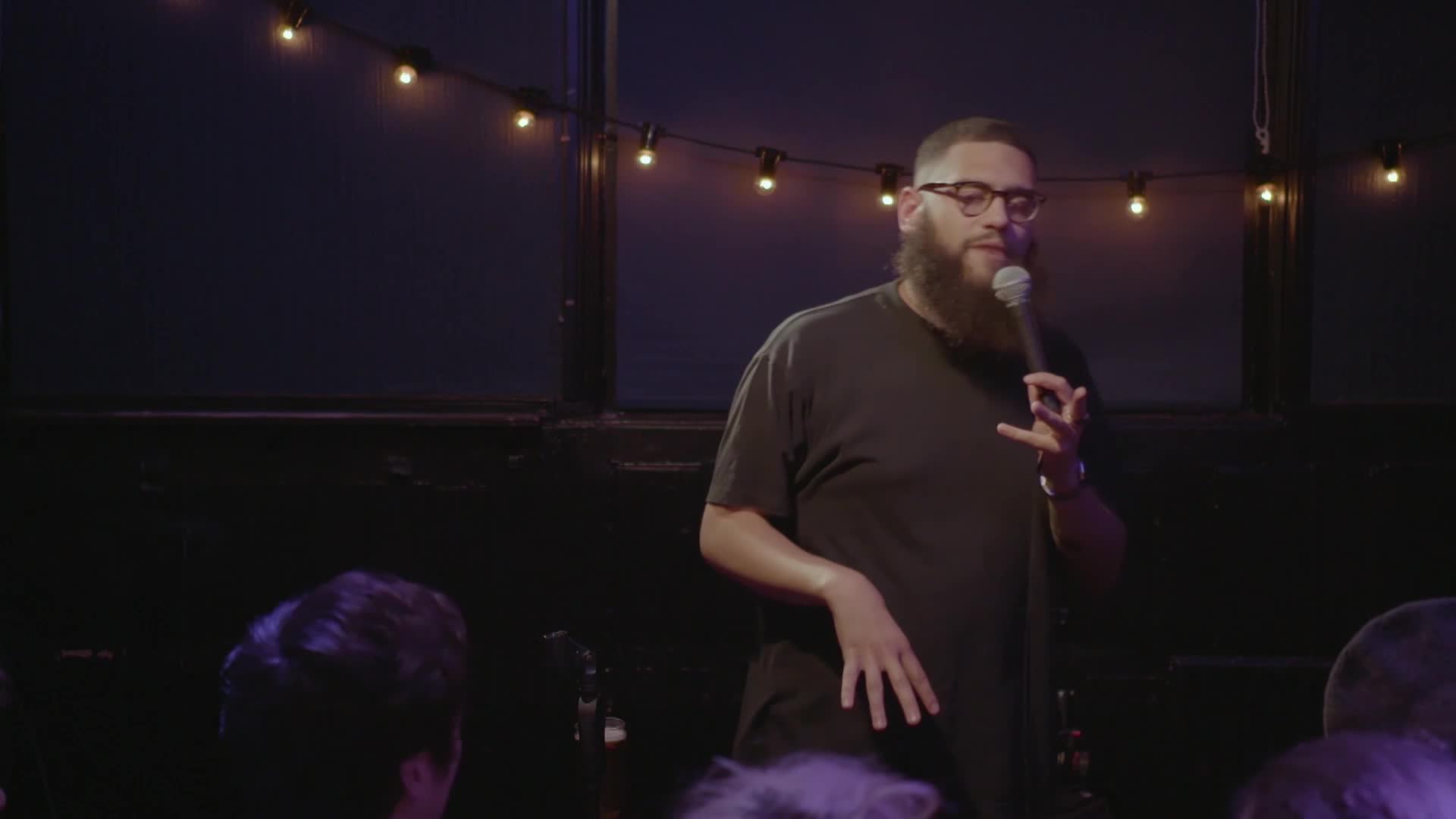 A man with a full beard and glasses stands on a stage, holding a microphone. He gestures with his hands as if he's telling a story.