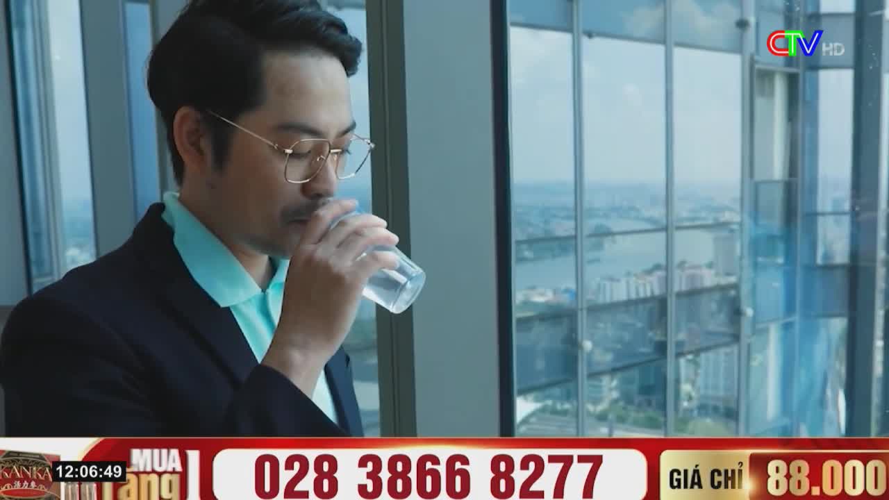 A man in a suit and glasses takes a drink from a clear glass, the city visible through the window behind him. The Ca Mau TV logo is visible in the corner.