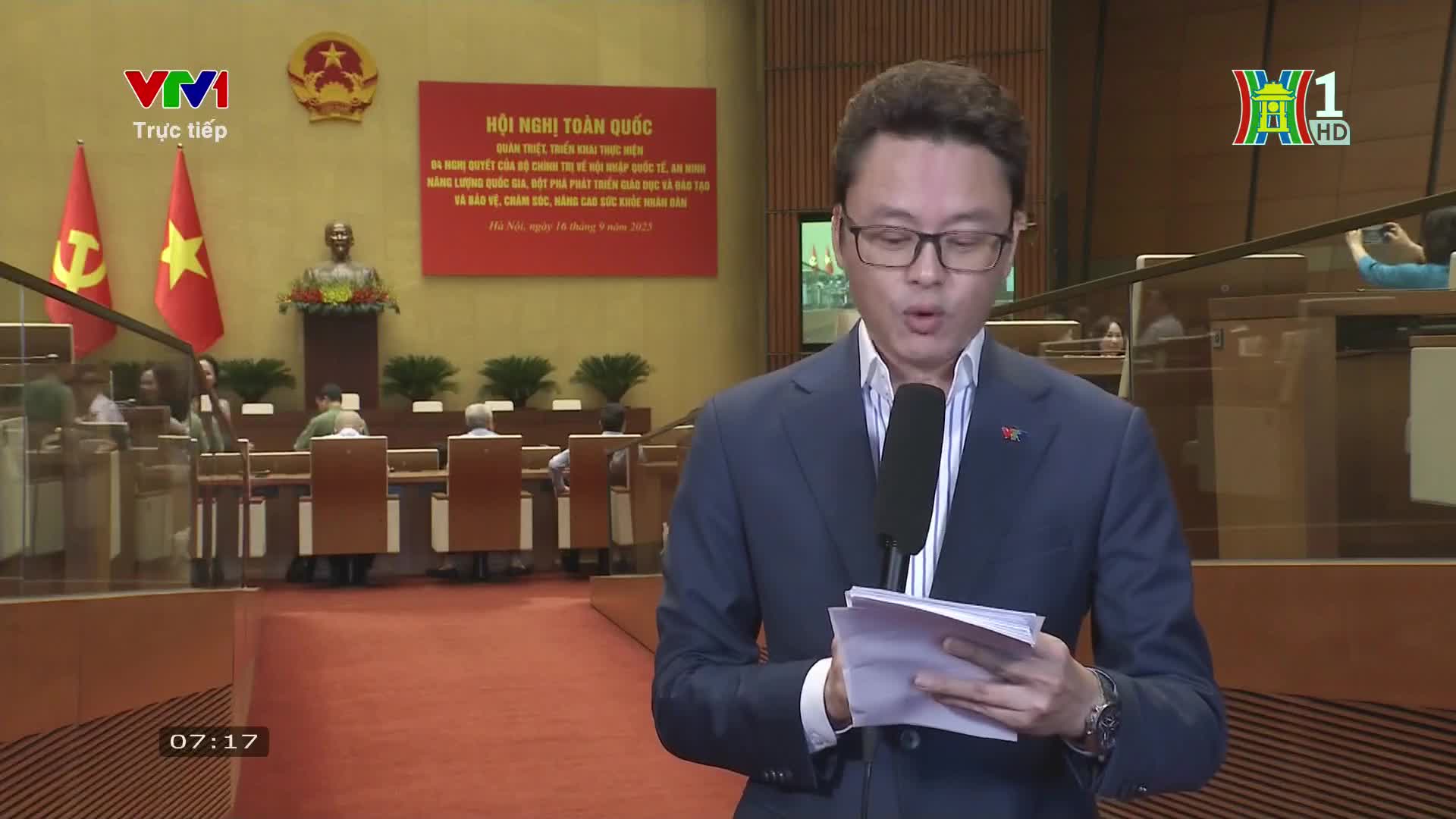 A man in a suit speaks into a microphone, reading from a stack of papers. Behind him, a large room is set up for a conference, with flags and a bust of a lea...