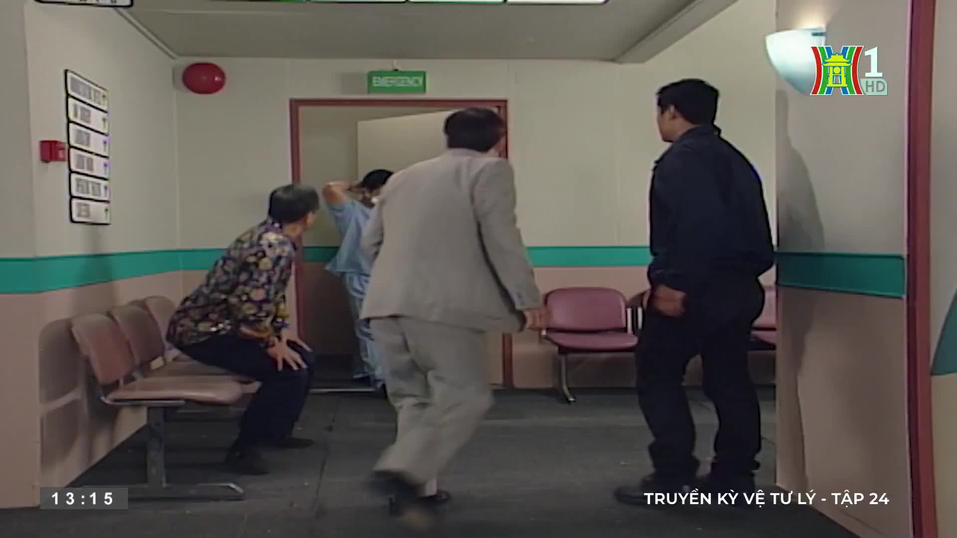 A man in a grey suit rushes forward, while a guard in black stands still. An older woman sits on a bench, as a doctor in blue scrubs exits a doorway.