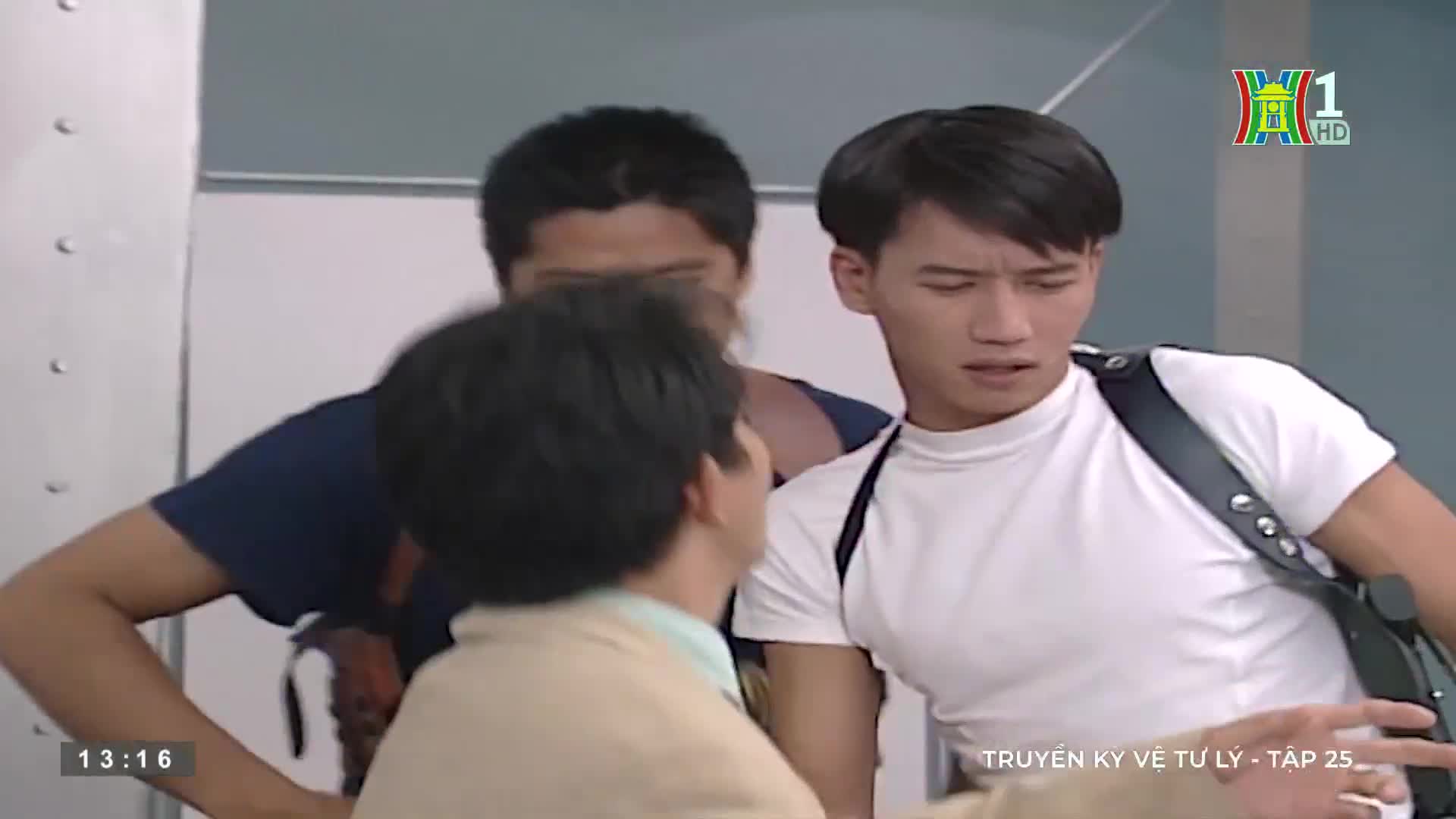 A man in a white t-shirt looks concerned, his eyes wide as he listens to someone off-screen. Two other men stand behind him, one with his arm resting on the ...