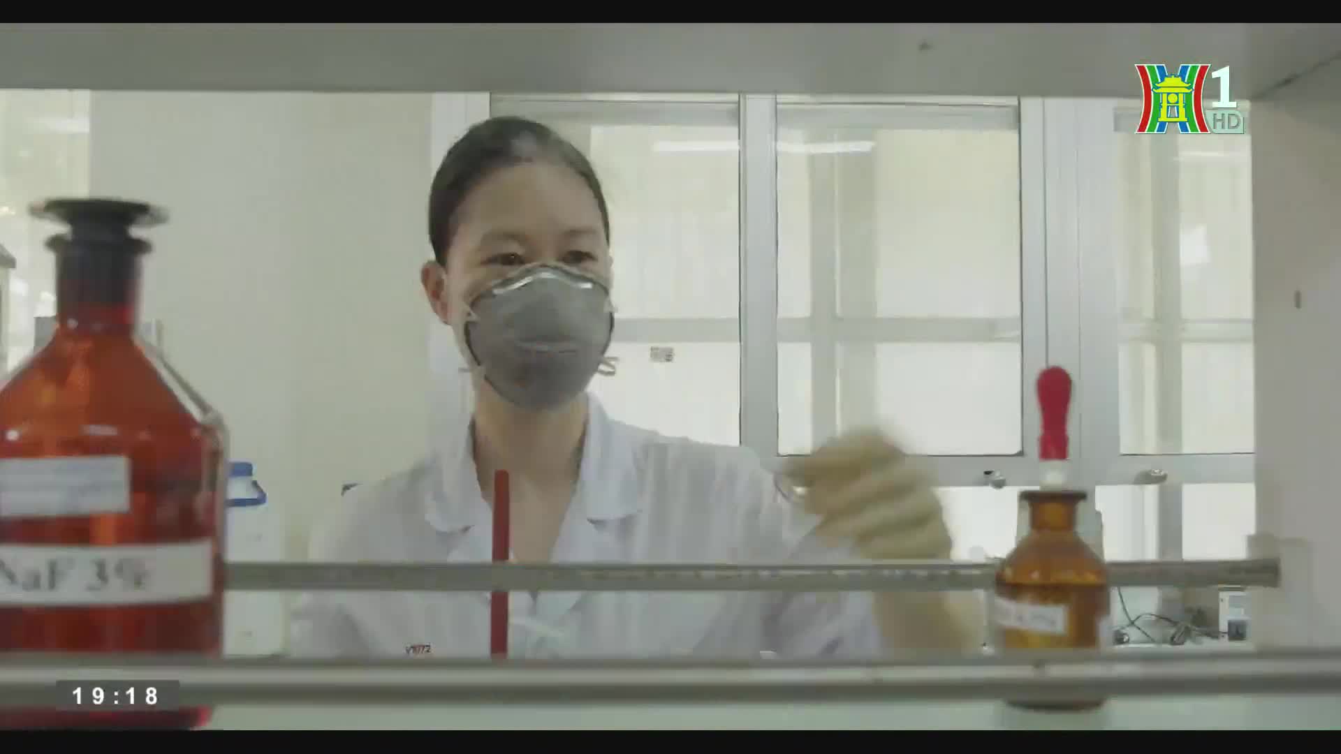 A woman in a lab coat and mask works with equipment, her gloved hand moving quickly.  A bottle of NaF 3% sits on the counter, visible on HanoiTV1.