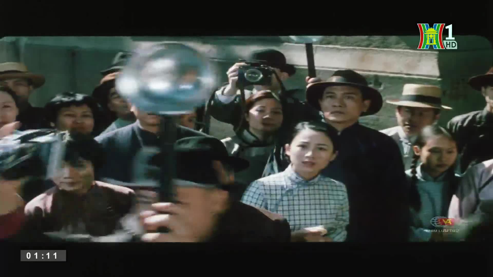 A crowd of people, many in hats, is gathered, some looking directly at the camera. A man holds a camera high, while another obscures the view with a large fl...