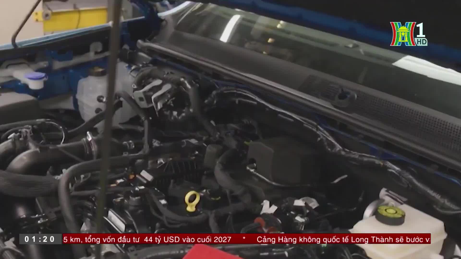 The open hood reveals the intricate engine of a blue vehicle, likely part of a segment from HanoiTV1. A yellow cap and various black tubes and wires are visi...