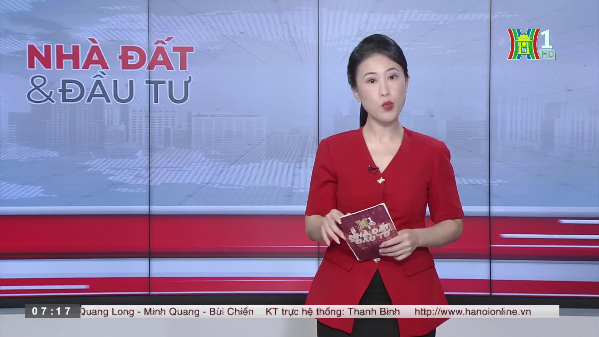 A woman in a red jacket stands in front of a backdrop, holding a small booklet. The HanoiTV1 logo is visible in the corner, and the screen behind her display...
