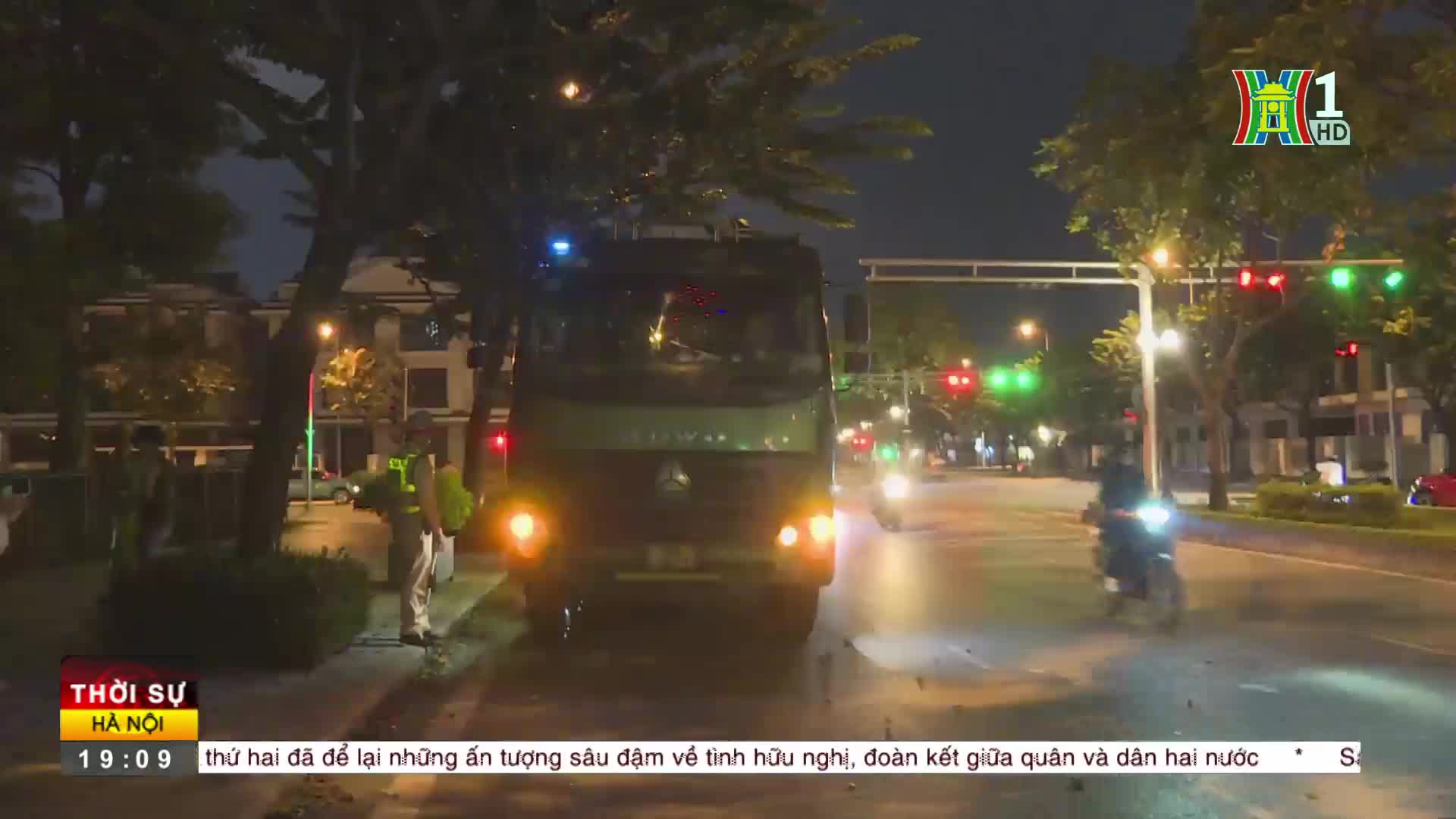 A large truck, its headlights on, is moving slowly down a street at night. The HanoiTV1 broadcast is showing a news report, with traffic lights visible in th...