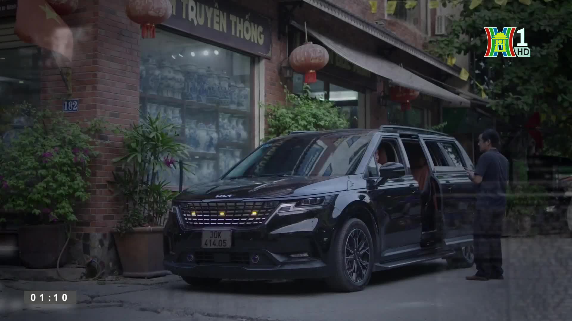 A black Kia minivan sits parked on a Hanoi street, its door open as a man stands nearby. The scene is framed by a shop with a sign in Vietnamese and a logo f...