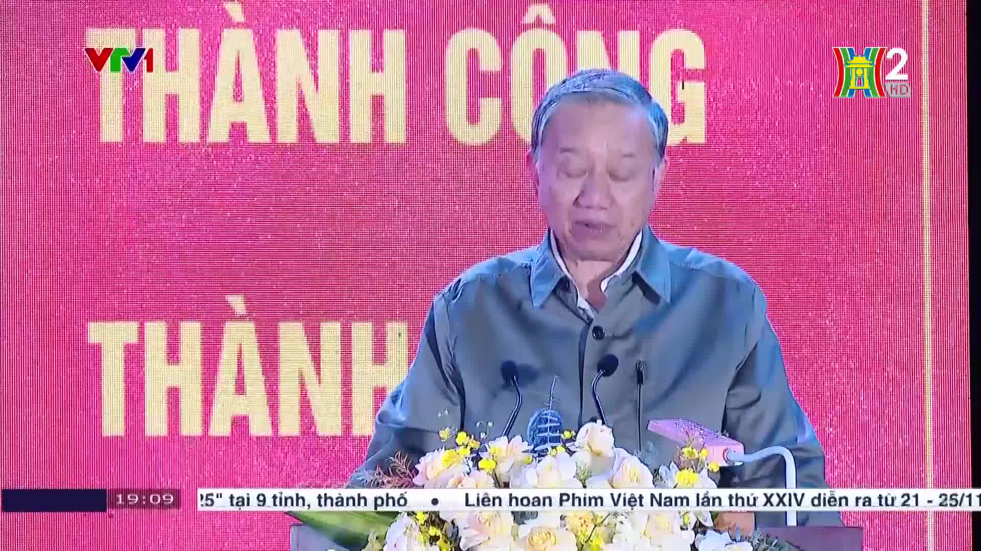 A man in a green shirt is speaking at a podium, with the Vietnam Television logo visible on the screen behind him. The broadcast is from HanoiTV2, and the ba...