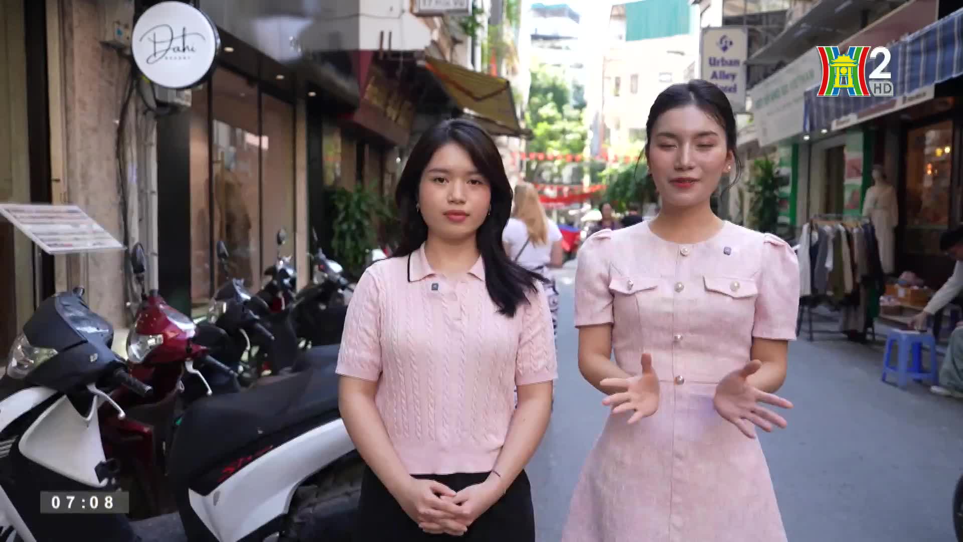 Two women in matching pink outfits stand on a narrow street, gesturing with their hands. Behind them, motorbikes line the sidewalk, and shops and pedestrians...