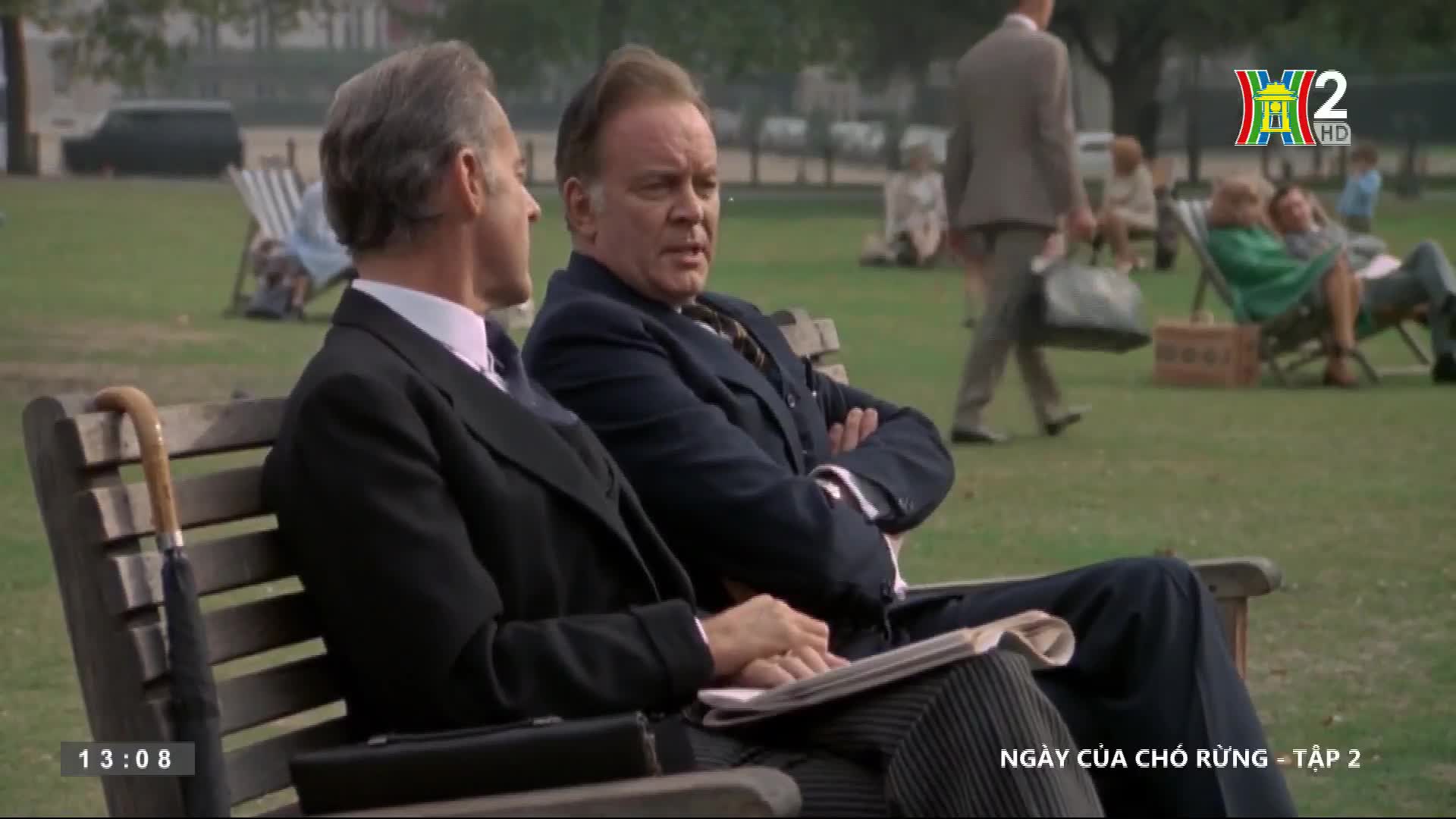 Two men in dark suits sit on a bench, one with arms crossed, the other holding a newspaper. People stroll across the green, and a couple relaxes in folding c...