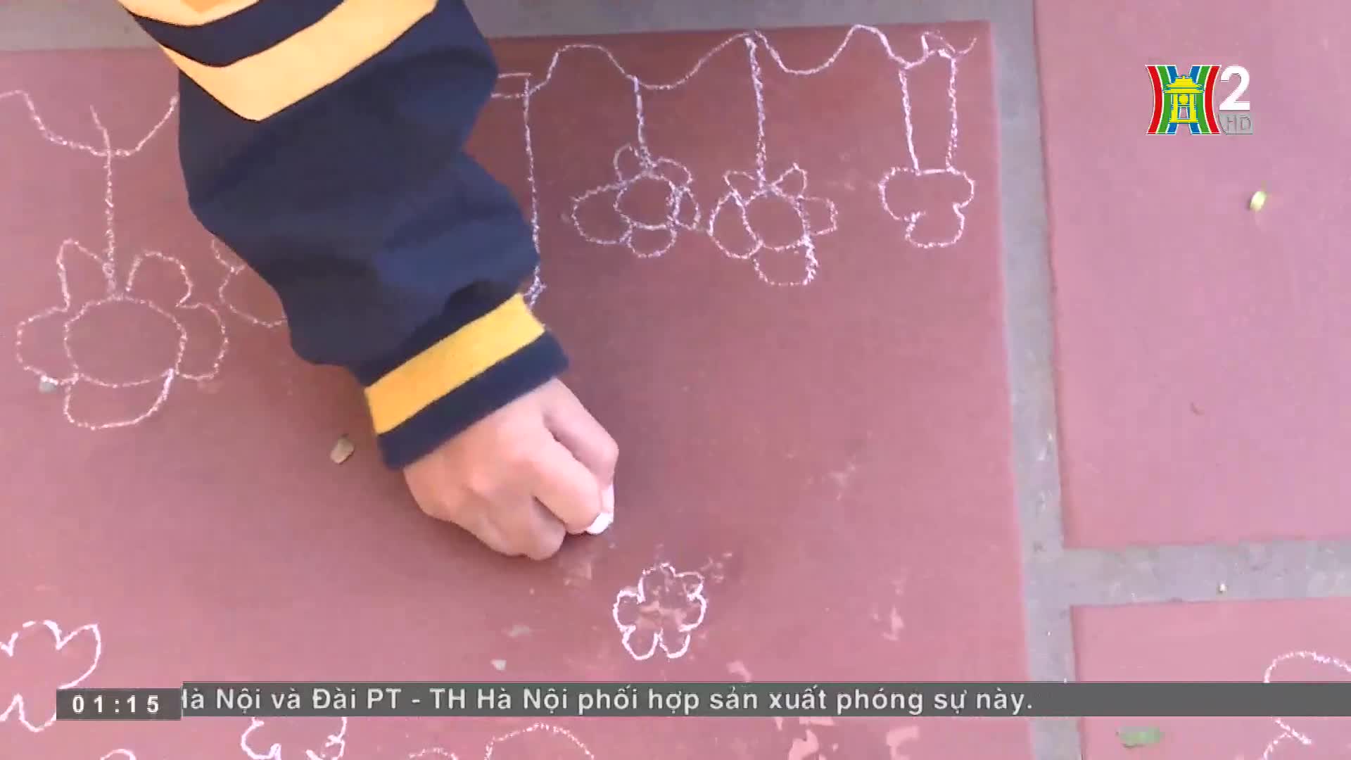 A hand, clad in a navy sleeve with yellow stripes, carefully draws white chalk flowers on a red surface. The HanoiTV2 logo is visible in the corner, and the ...