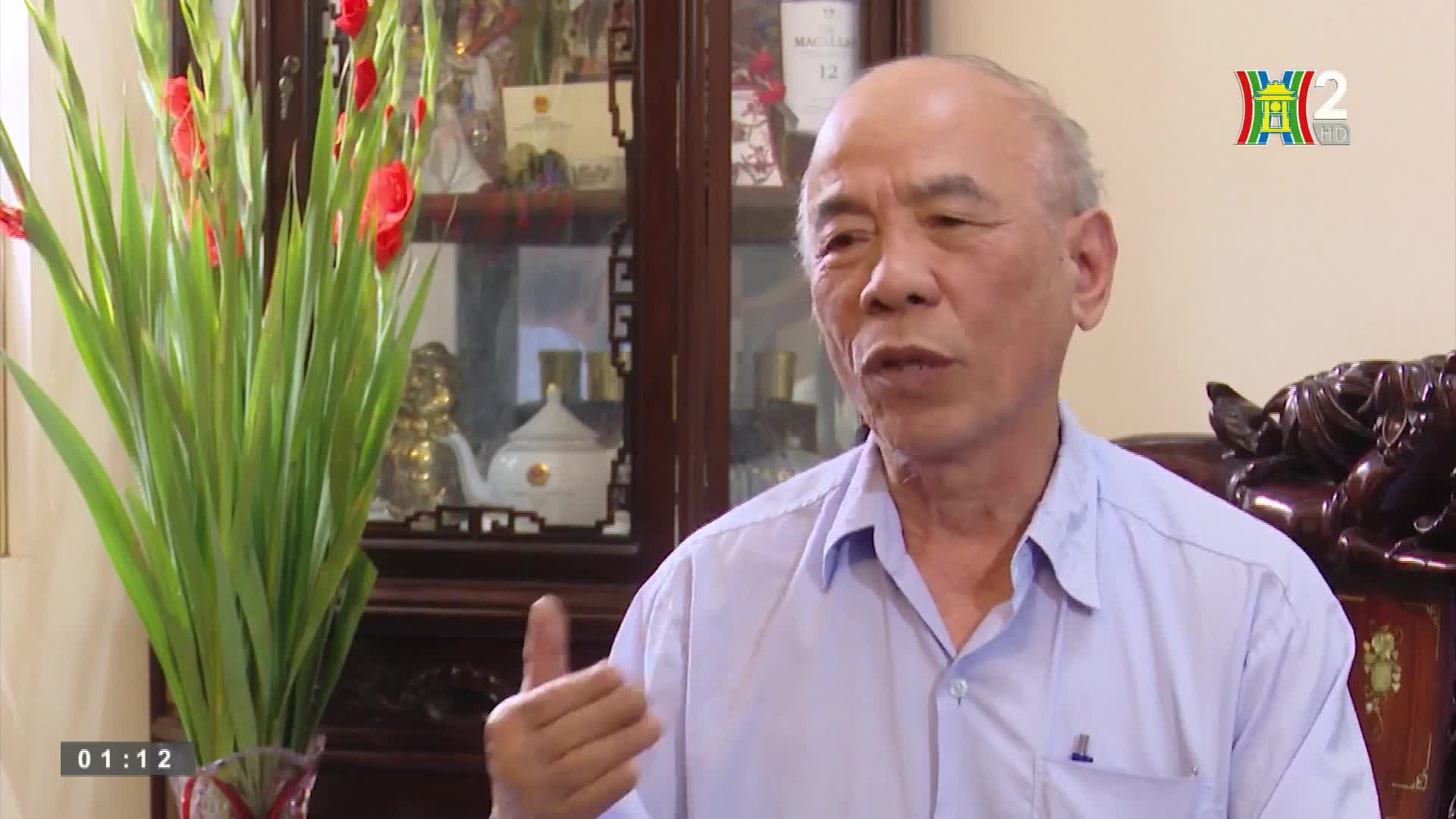 A man in a light blue shirt gestures with his right hand as he speaks. Behind him, a vase of red gladioli stands next to a glass cabinet, and the HanoiTV2 lo...