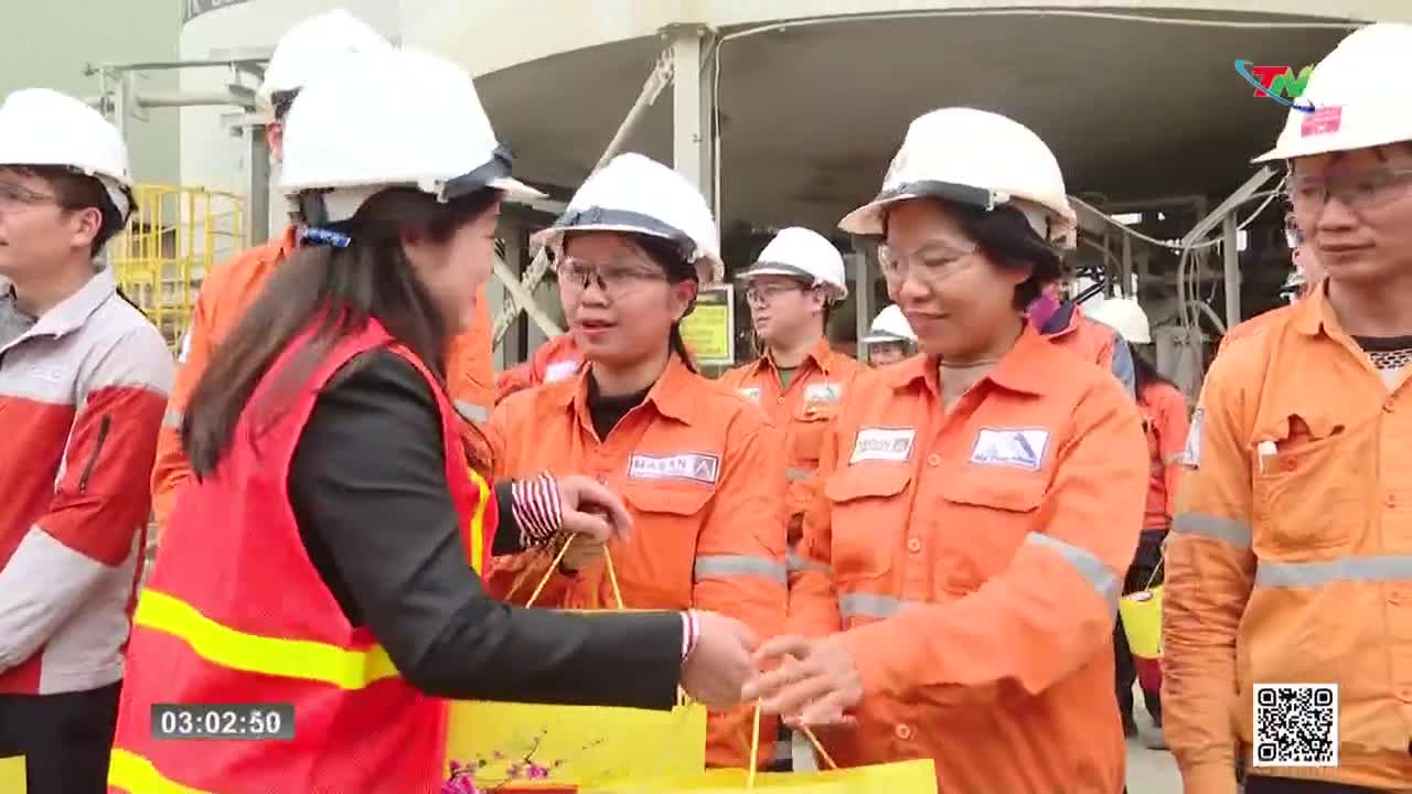 A woman in a red vest hands a yellow gift bag to another woman wearing an orange jumpsuit. Several other workers in similar attire stand nearby, their faces ...