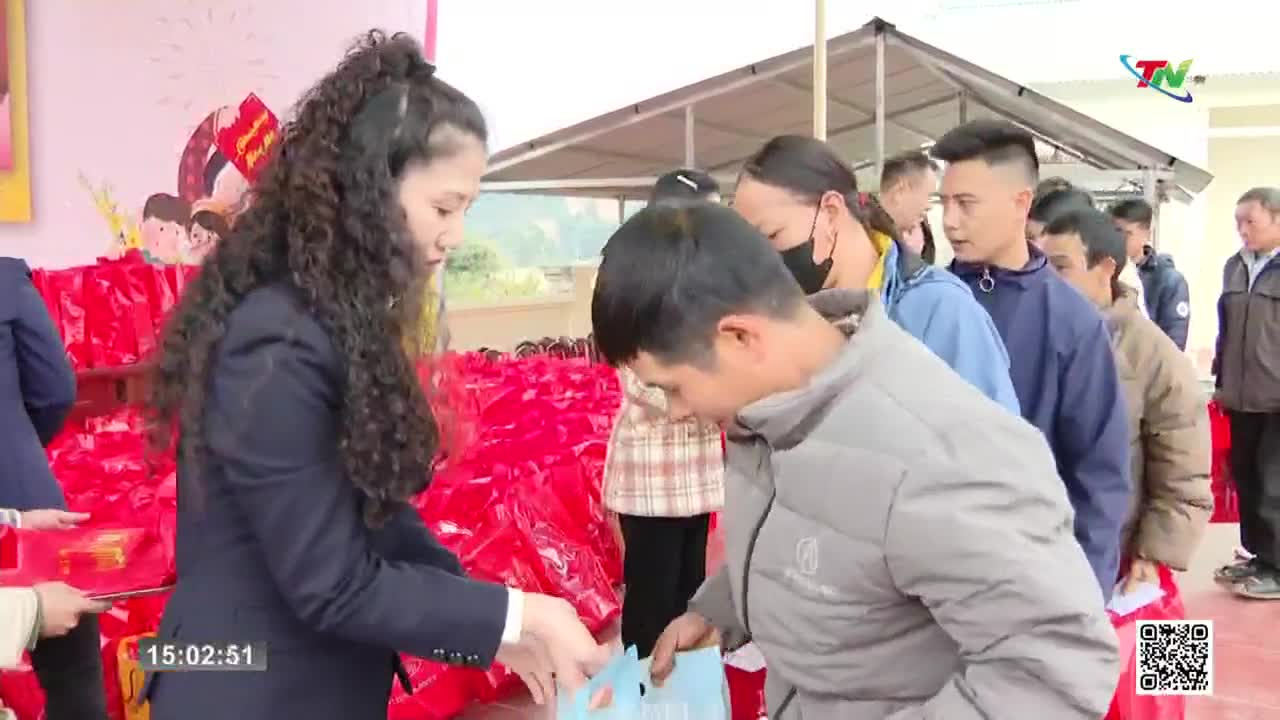 A woman in a dark suit hands a blue bag to a man in a gray jacket, while others wait in line behind him. The scene unfolds outdoors, with a backdrop of red b...